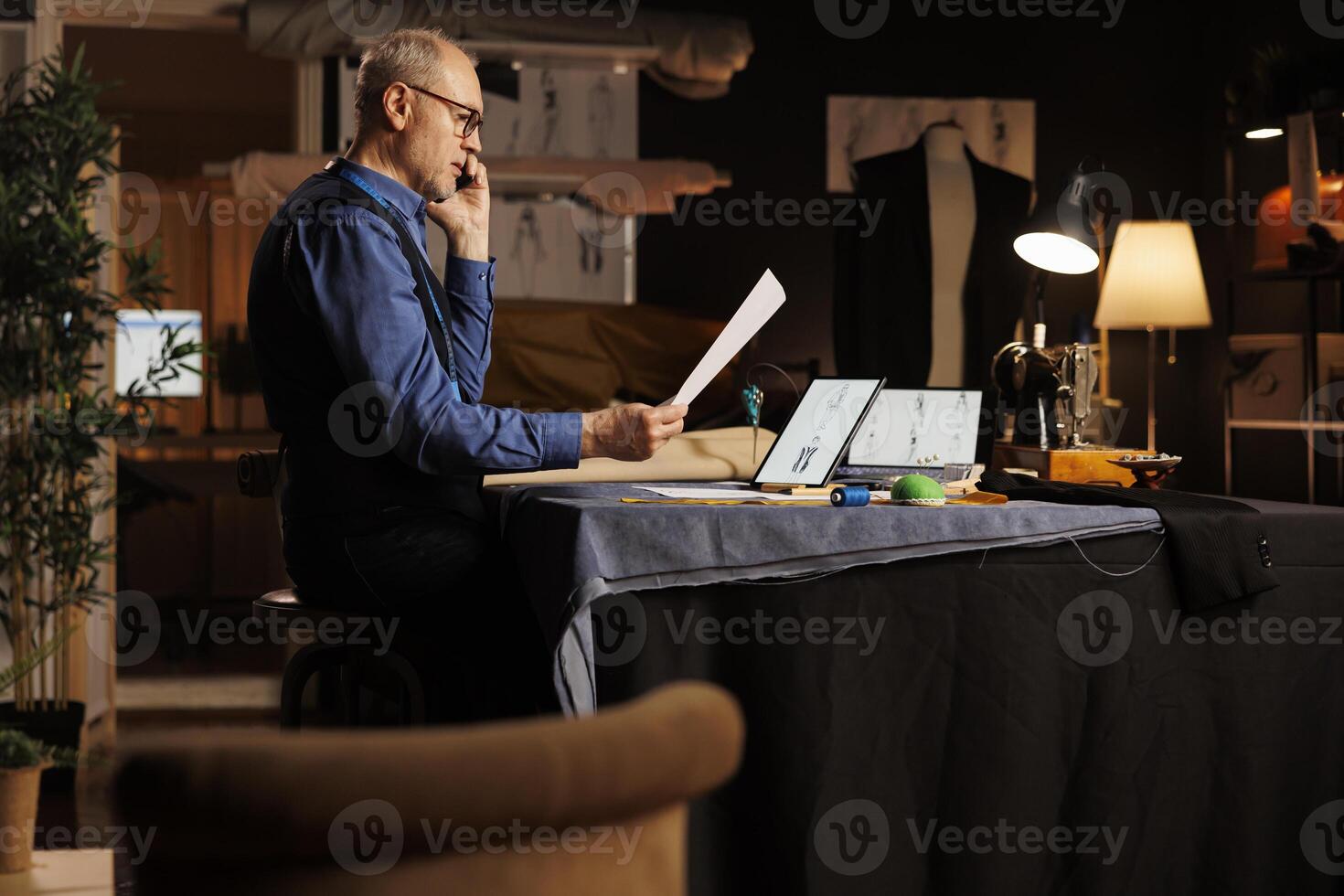 Elderly tailor talking over the phone with customer, asking for details about comissioned sartorial bespoken adjustments and customizations. Couturer in atelier on call with client photo