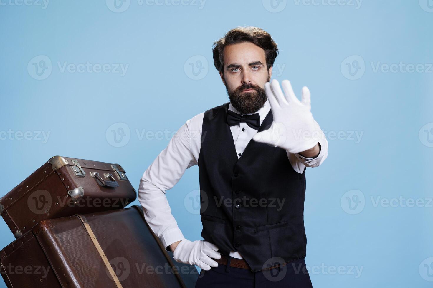 Professional doorman shows palm with gloves to express rejection over blue background, creating advertisement with refusal symbol. Bellhop showcasing his disagreement to ideas, dismissal. photo