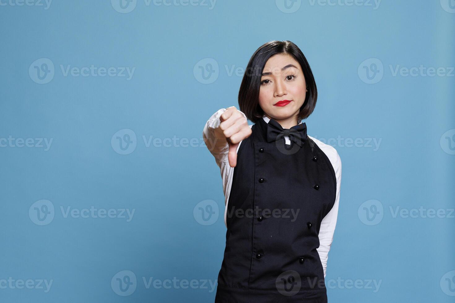 Disappointed asian waitress showing thumb down gesture, symbolizing catering service dissatisfaction and disapproval portrait. Young woman receptionist showcasing dislike symbol photo