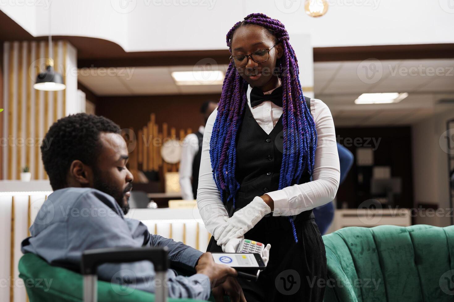 African American woman waitress giving pos machine to hotel guest waiting in lobby for check-in procedure. Tourist holding smartphone paying for order using nfc technology and banking app photo