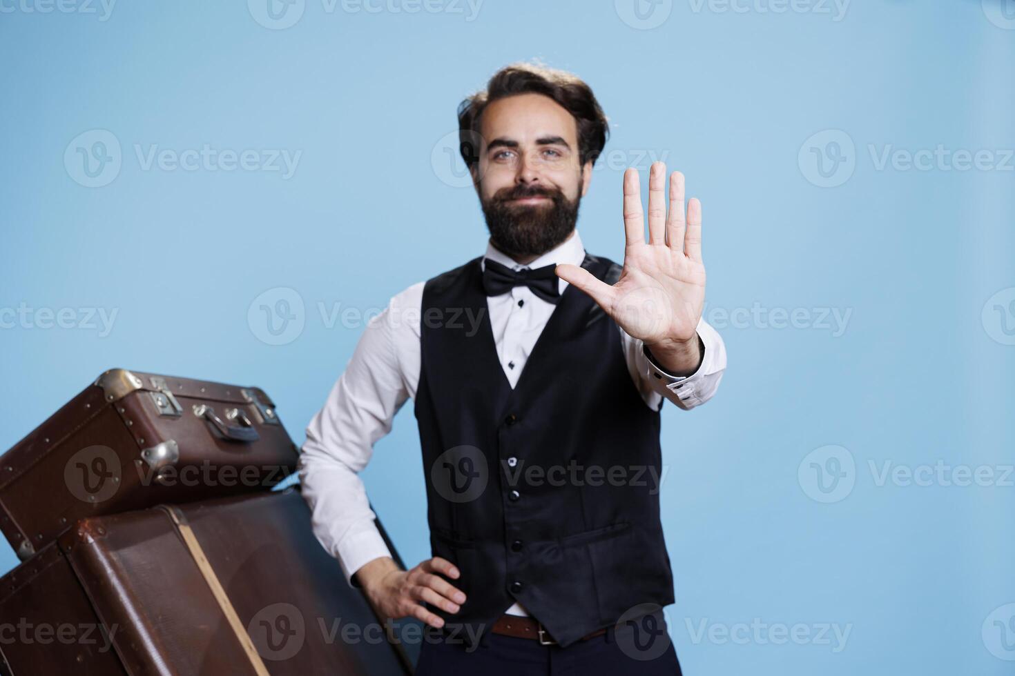 Employee showing stop sign on camera, hotel concierge in suit and tie implying refusal and rejection with hand raised in studio. Professional bellboy standing against blue background. photo