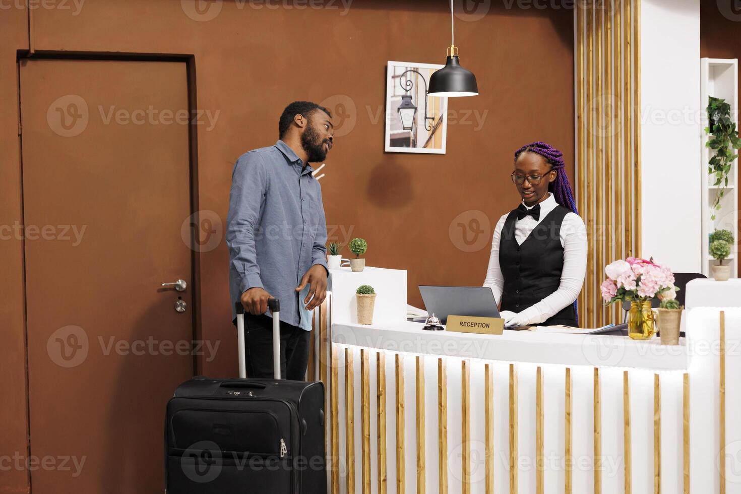 Friendly African American woman receptionist using laptop computer while standing behind front desk, talking with hotel guest. Black guy tourist checking in at resort. Resort holidays photo