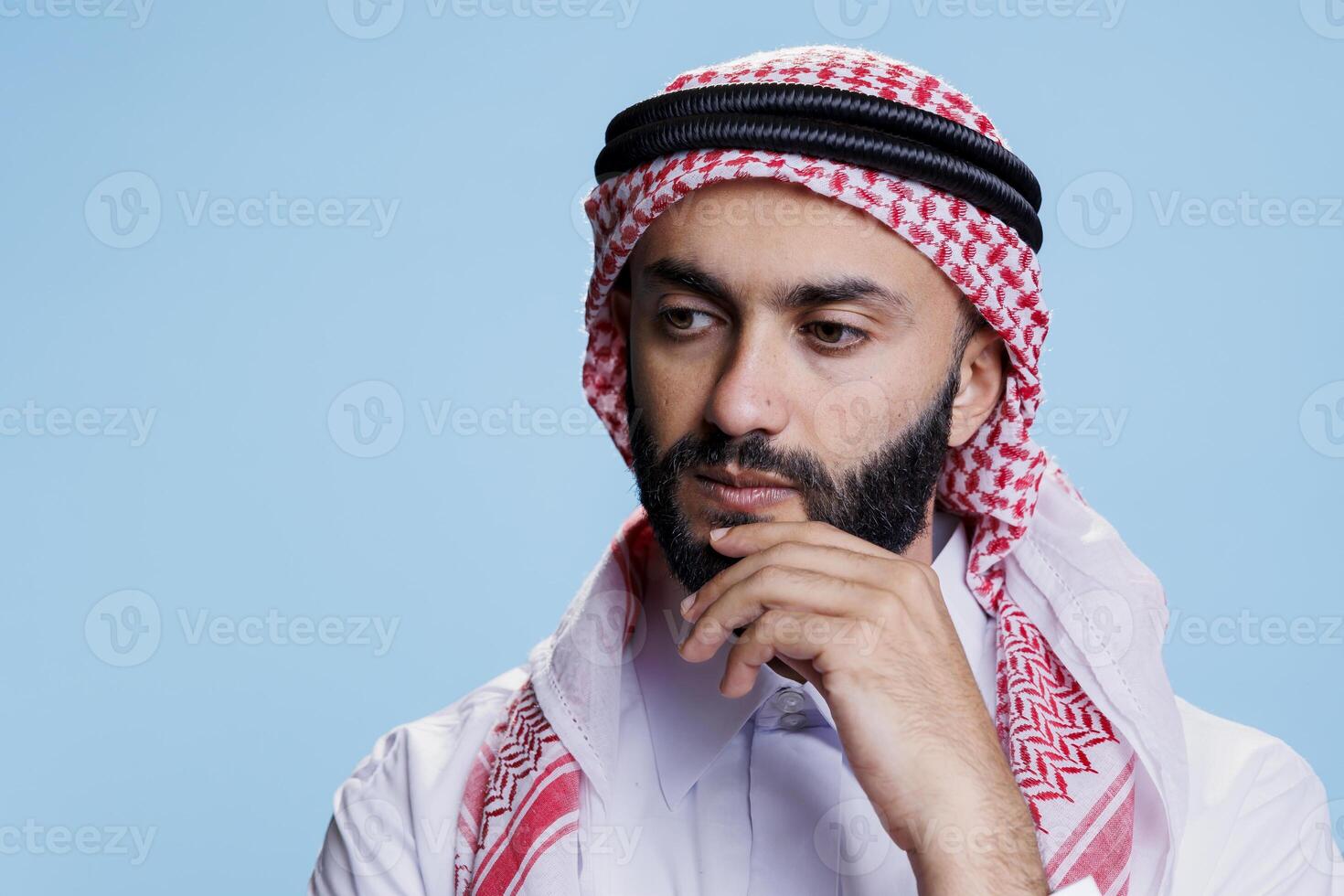 Thoughtful man wearing traditional muslim clothes rubbing chin and looking away. Arab person dressed in checkered ghutra headscarf standing in contemplation in studio on blue background photo