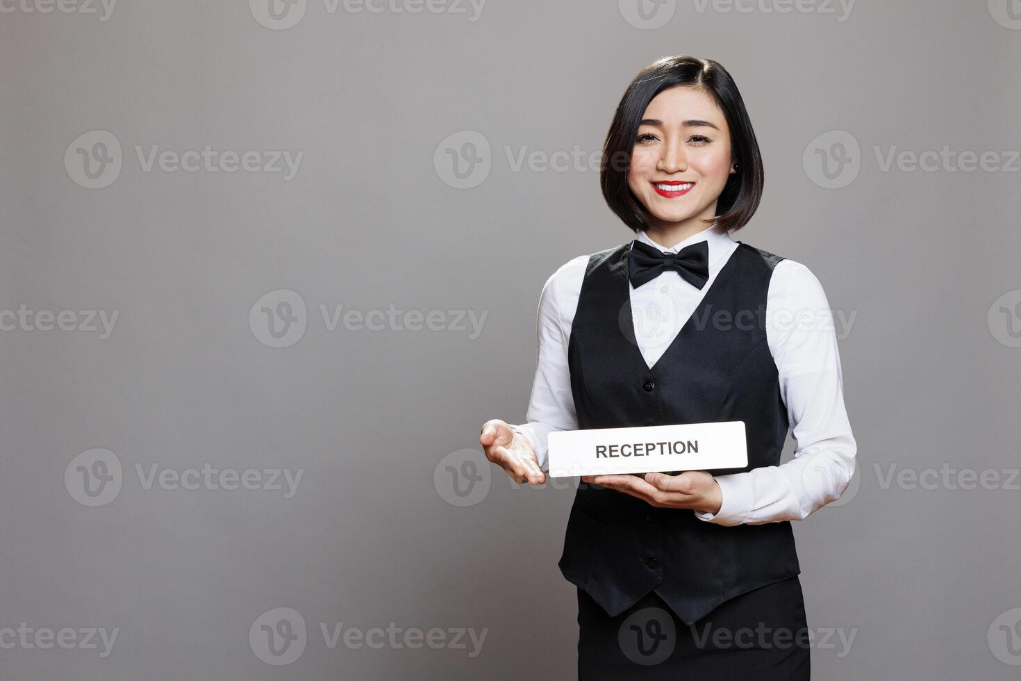 Smiling asian receptionist holding reception plate and looking at camera with cheerful face expression. Hospitality service employee in uniform showing hotel role signage portrait photo