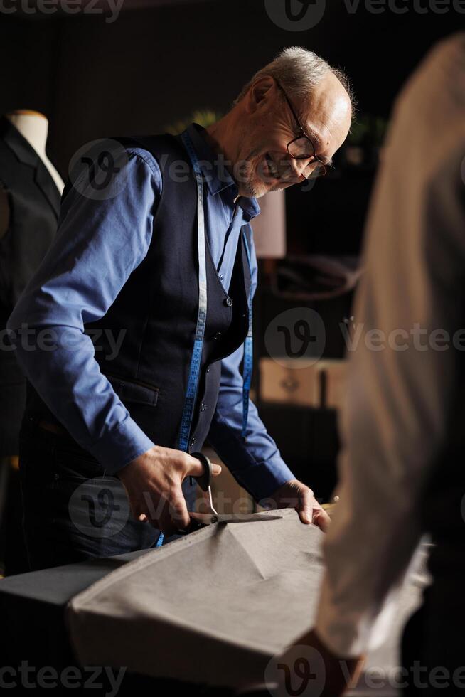 Happy couturiers in tailoring studio manufacturing premium quality sartorial garments. Senior suitmaker showing apprentice how to masterfully cut fabric and make alterations photo