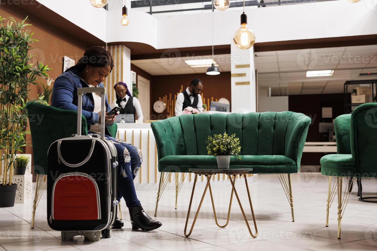 African American woman traveler sitting in hotel with luggage, using smartphone texting message while waiting for check-in in lobby. Female guest checking in via mobile device photo