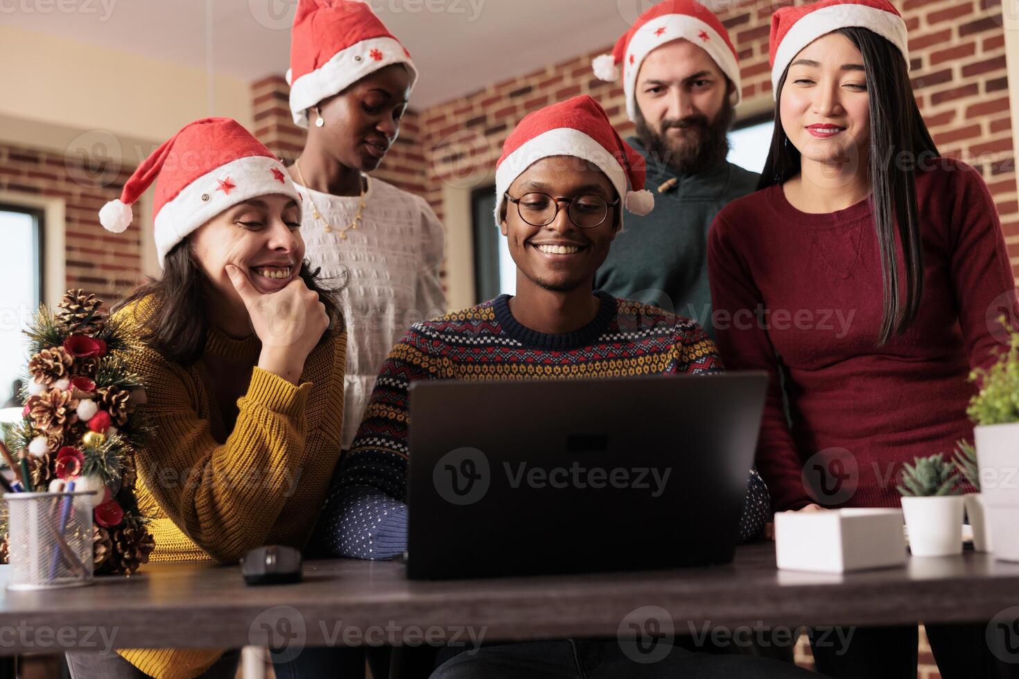Smiling workers helping african american man colleague to finish task before deadline on xmas eve. Cheerful diverse coworkers in santa hats teamwork during winter seasonal holiday in office photo