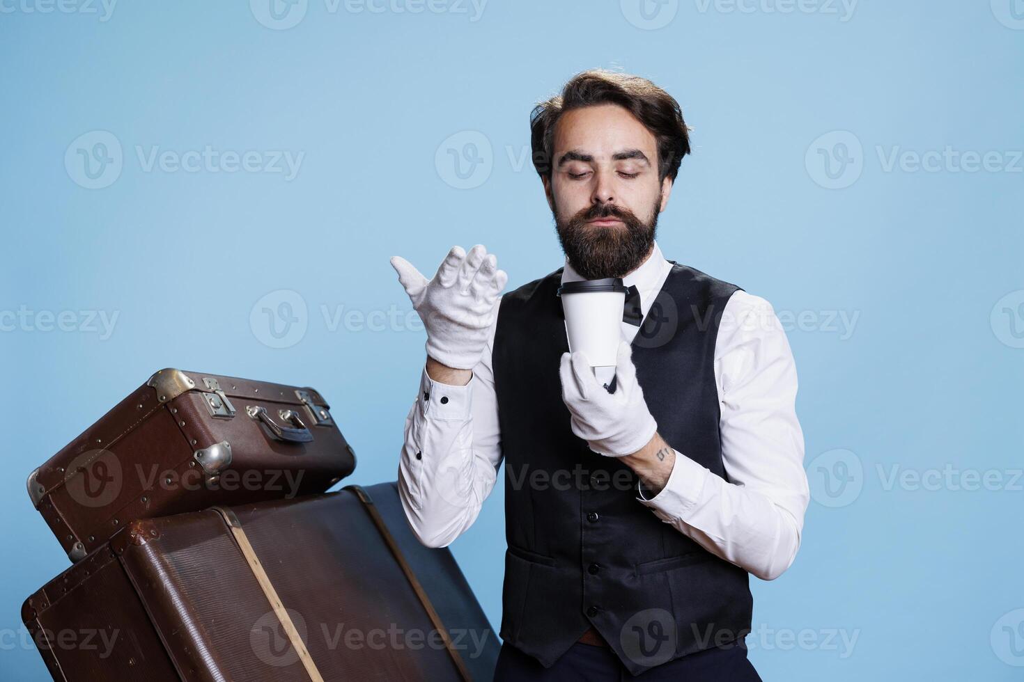 Elegant bellboy savouring smell of drink against blue background in studio, posing with cup of coffee on camera. Professional hotel concierge worker smells beverage with great aroma. photo