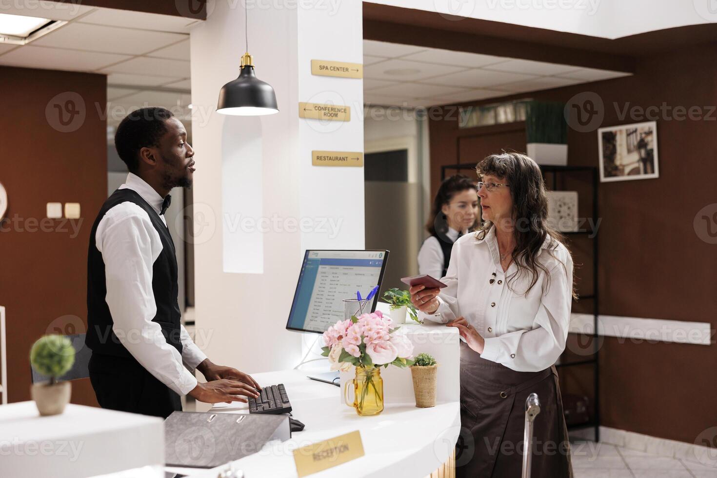 Arrival in hotel reception, elderly female customer checks in with receptionist assistance at front desk. African american concierge receives personal document from old woman for smooth registration. photo