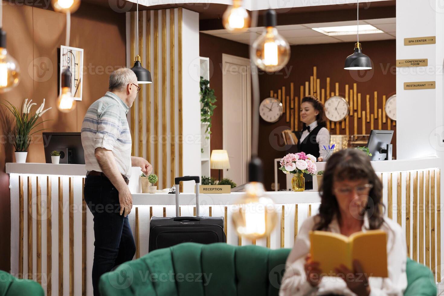 Elderly man visitor comes at front desk of hotel lobby and greeted by pleasant female receptionist. While waiting for check-in, senior woman sits on comfy sofa and reads a book in cozy lounge area. photo