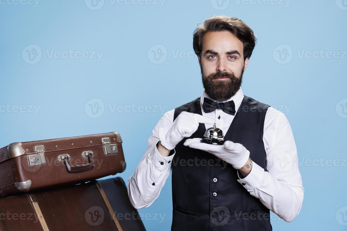 Bellboy with gloves holding service bell in studio, posing as hotel concierge employee to give assistance with luggage. Hotel worker in formal suit prepared to help guests, hospitality concept. photo