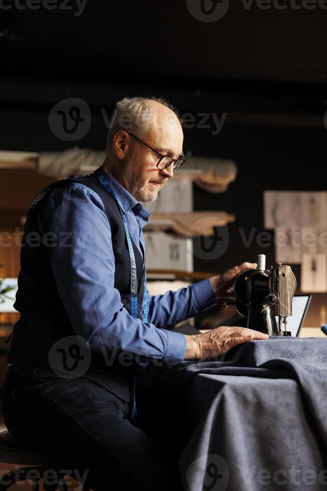 Senior tailor meticulously cutting fabric material with professional sewing machine in atelier shop workshop. Skilled elderly suitmaker working on sartorial garments for upcoming customer comission photo