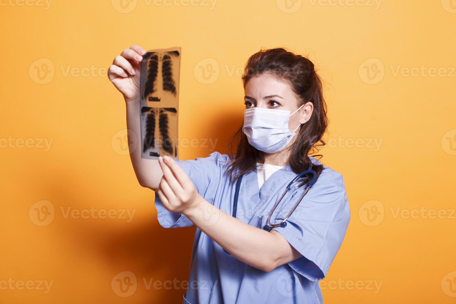 Medical practitioner with face mask analyzes radiography to find diagnosis. Assistant doctor wearing protection against virus and blue scrubs with stethoscope while looking at x ray scan in studio. photo