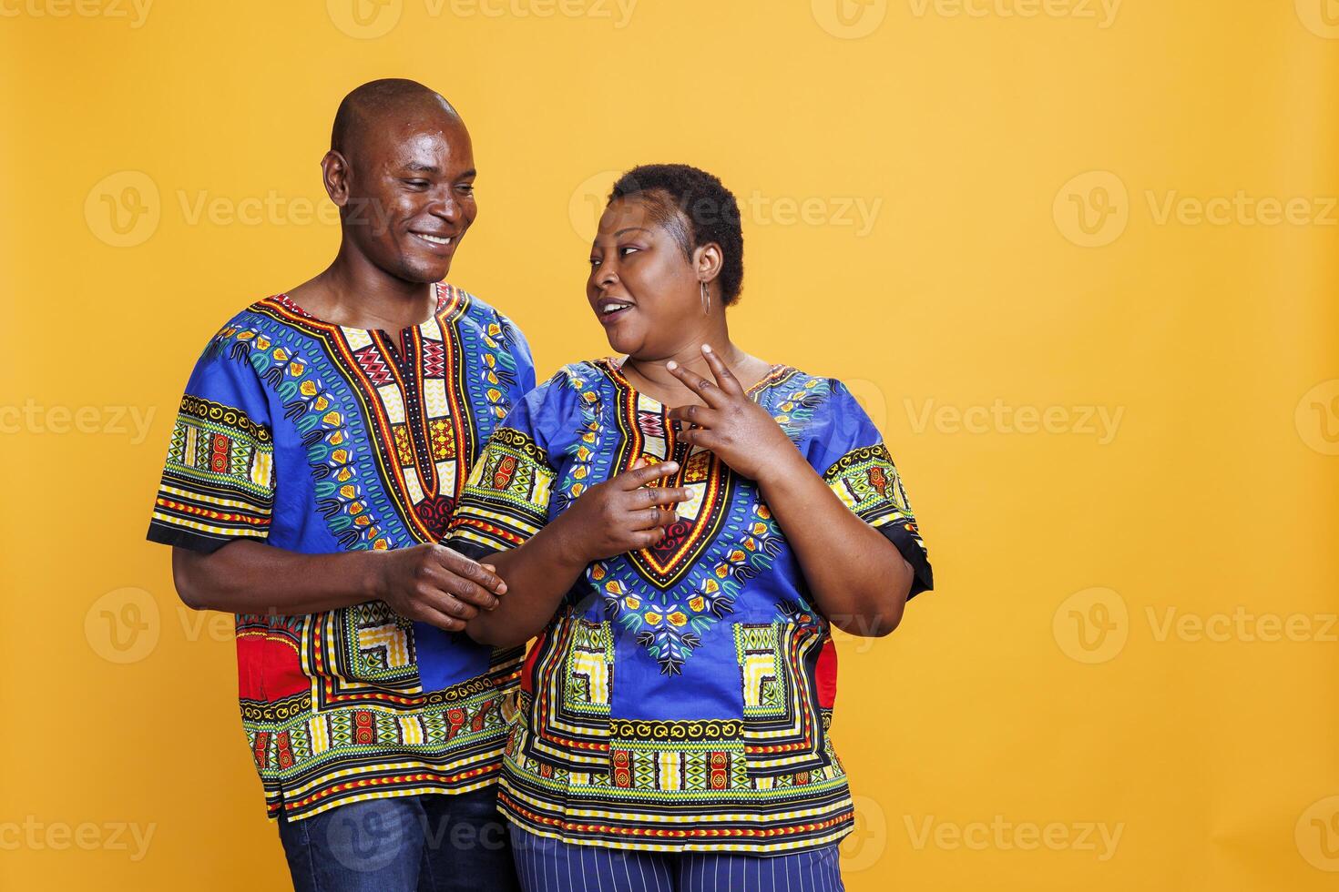 Smiling man and woman romantic pair chatting while standing in studio. Happy wife and husband wearing ethnic african american clothes talking and posing on orange background photo