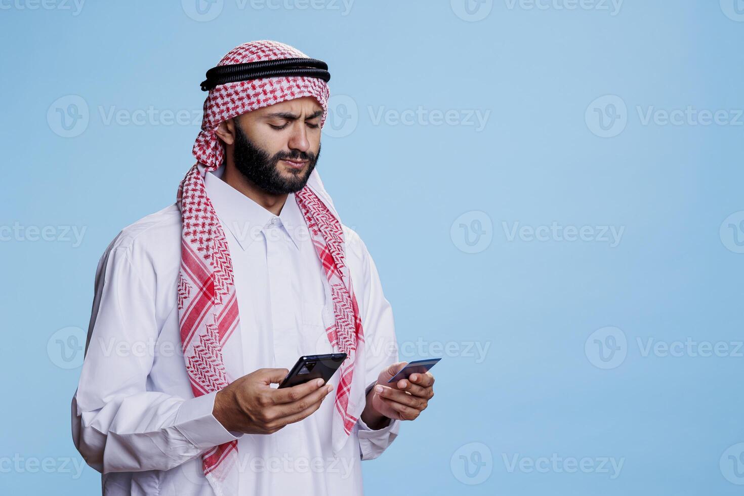 Muslim man in traditional clothes holding credit card and smartphone while shopping online. Arab with mobile phone making purchase in internet store using electronic banking photo