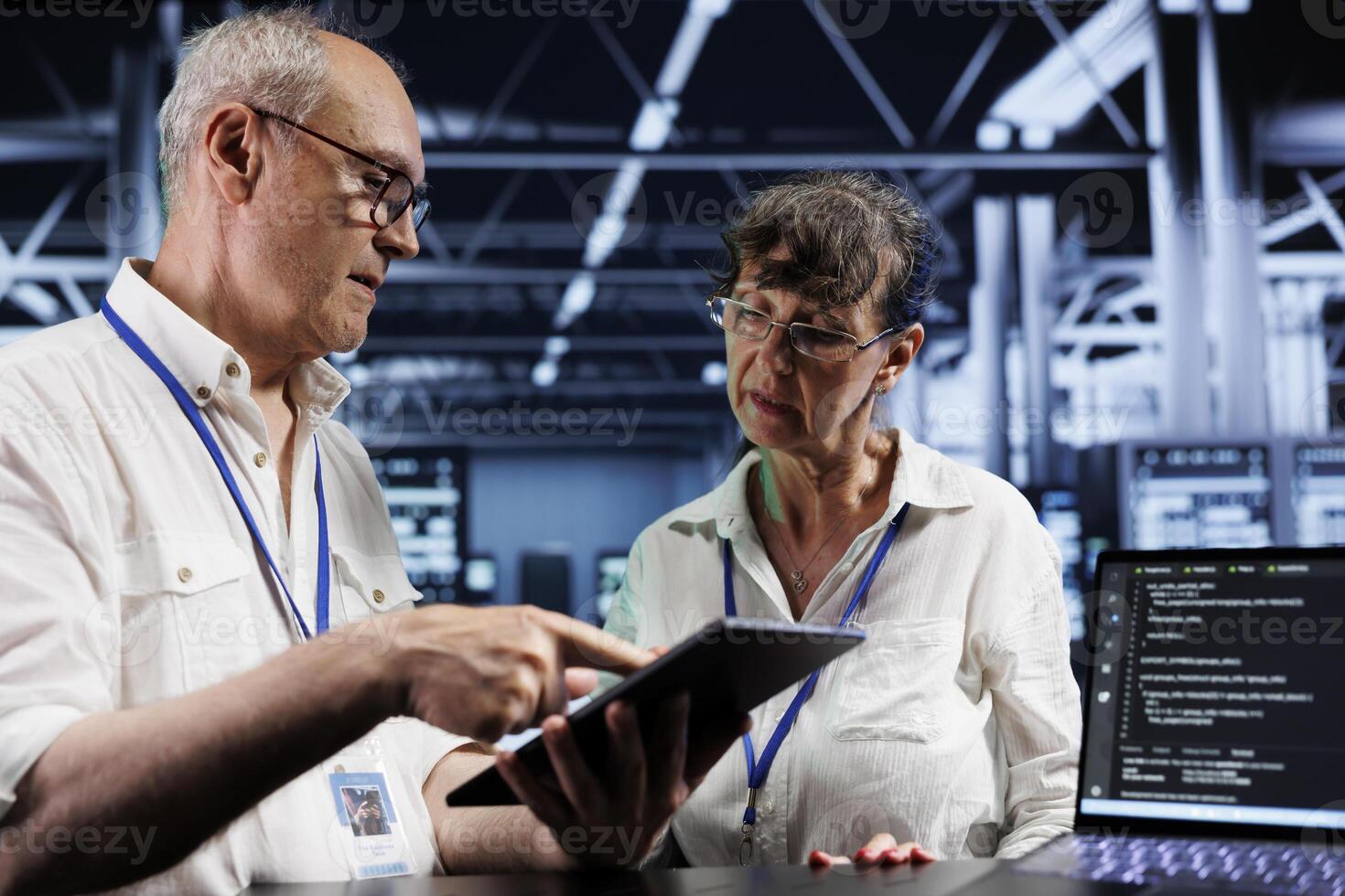 Teamworking colleagues updating server racks, writing intricate binary code scripts on tablet and laptop. Elderly coworkers using programming to fortify cybersecurity in high tech workspace photo