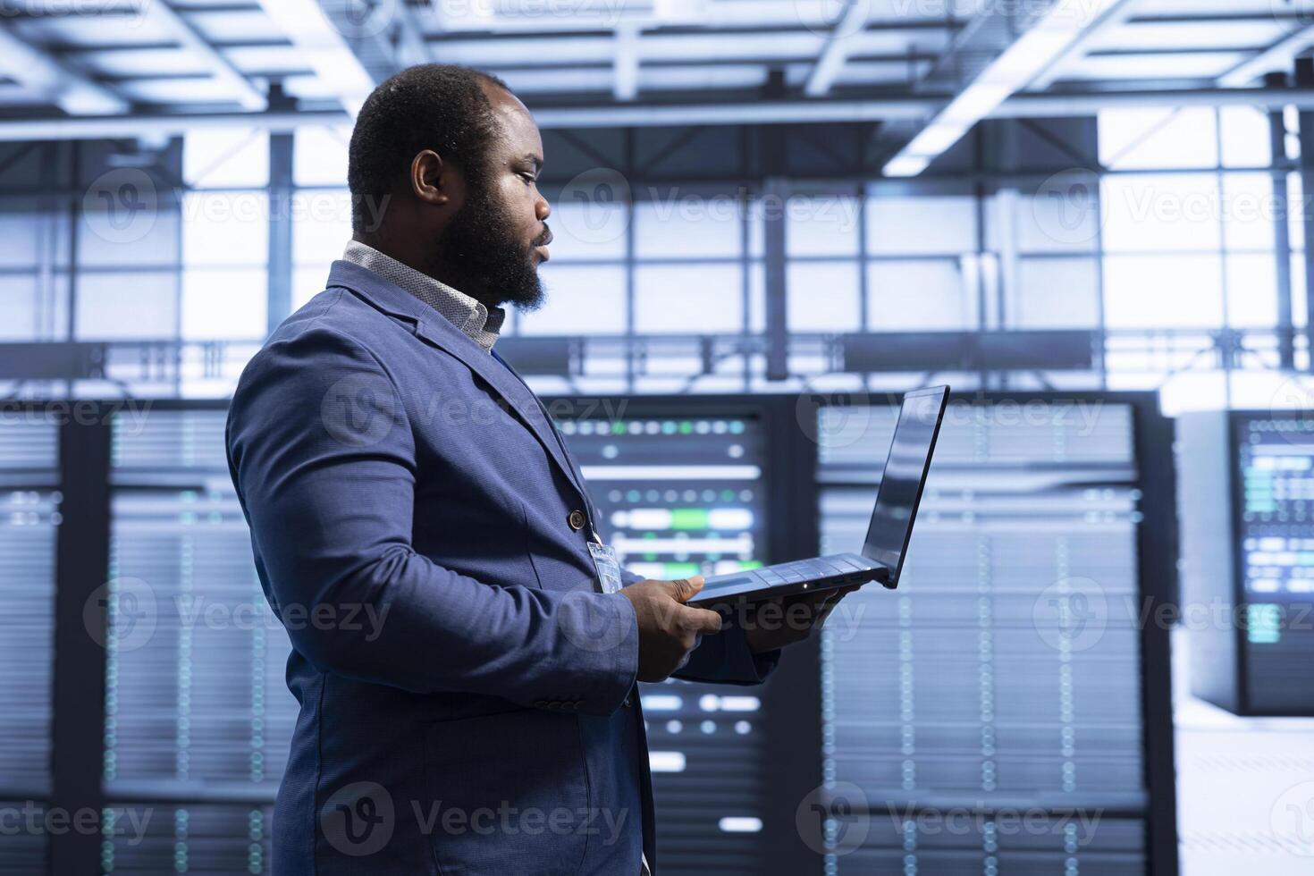 Software developer walking in data center, using laptop to configure network infrastructure such as routers, switches and firewalls. African american man in server farm looking for technical issues photo