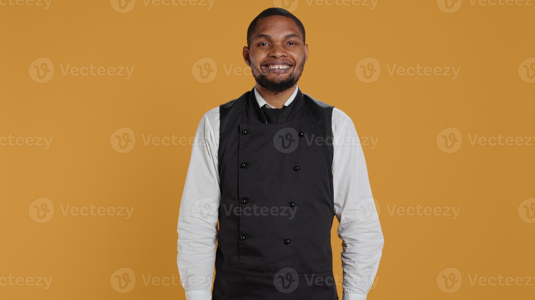 Waiter in apron and uniform showing a heart shape sign in studio, expressing his sincere feelings and true love. Dining employee presents a romantic gesture, sweet confident man. Camera B. photo