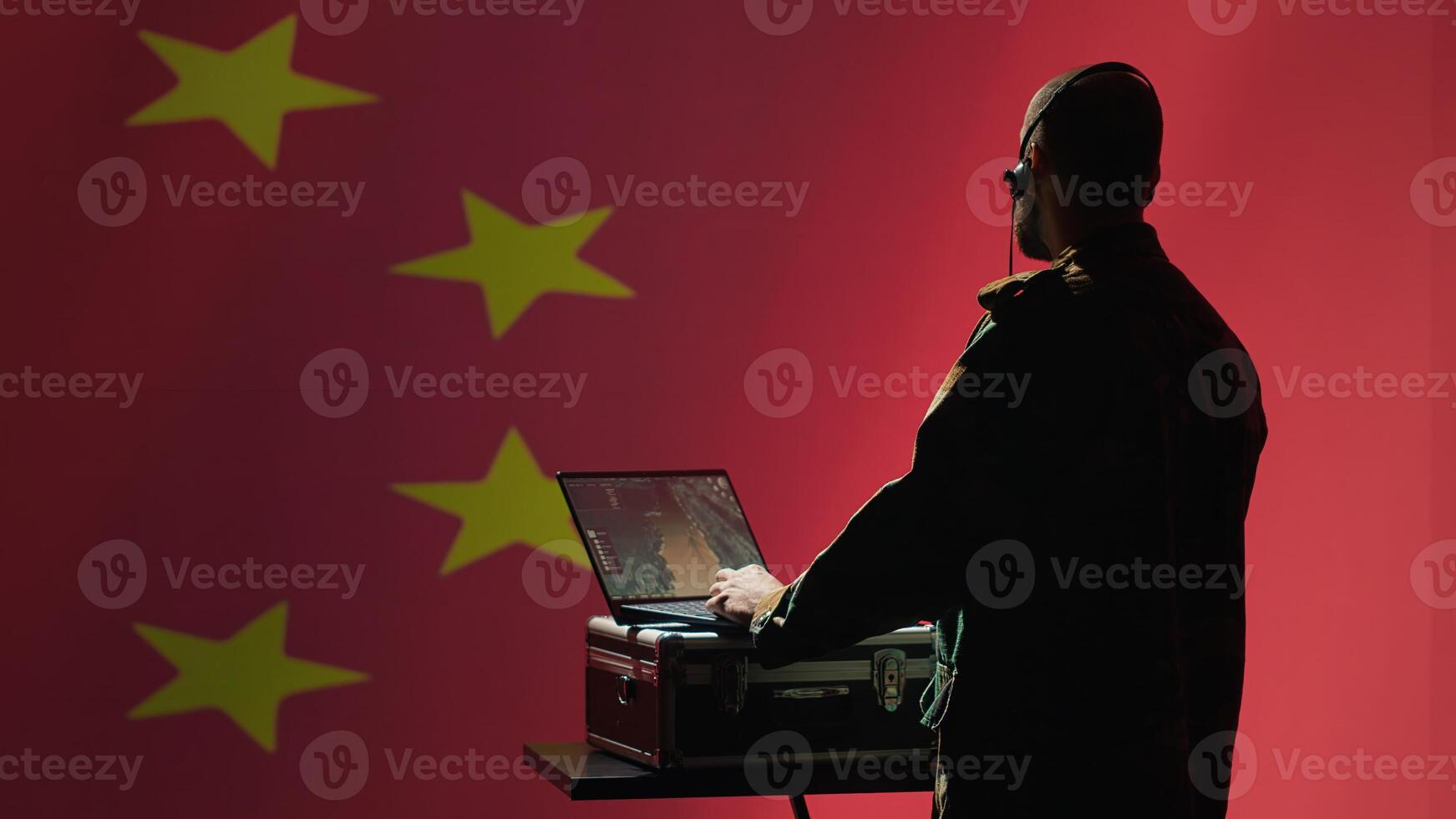 Chinese army military dispatcher looking at surveillance transmission on laptop, relaying informations for troops using headset, isolated over fluttering China flag background, camera A photo