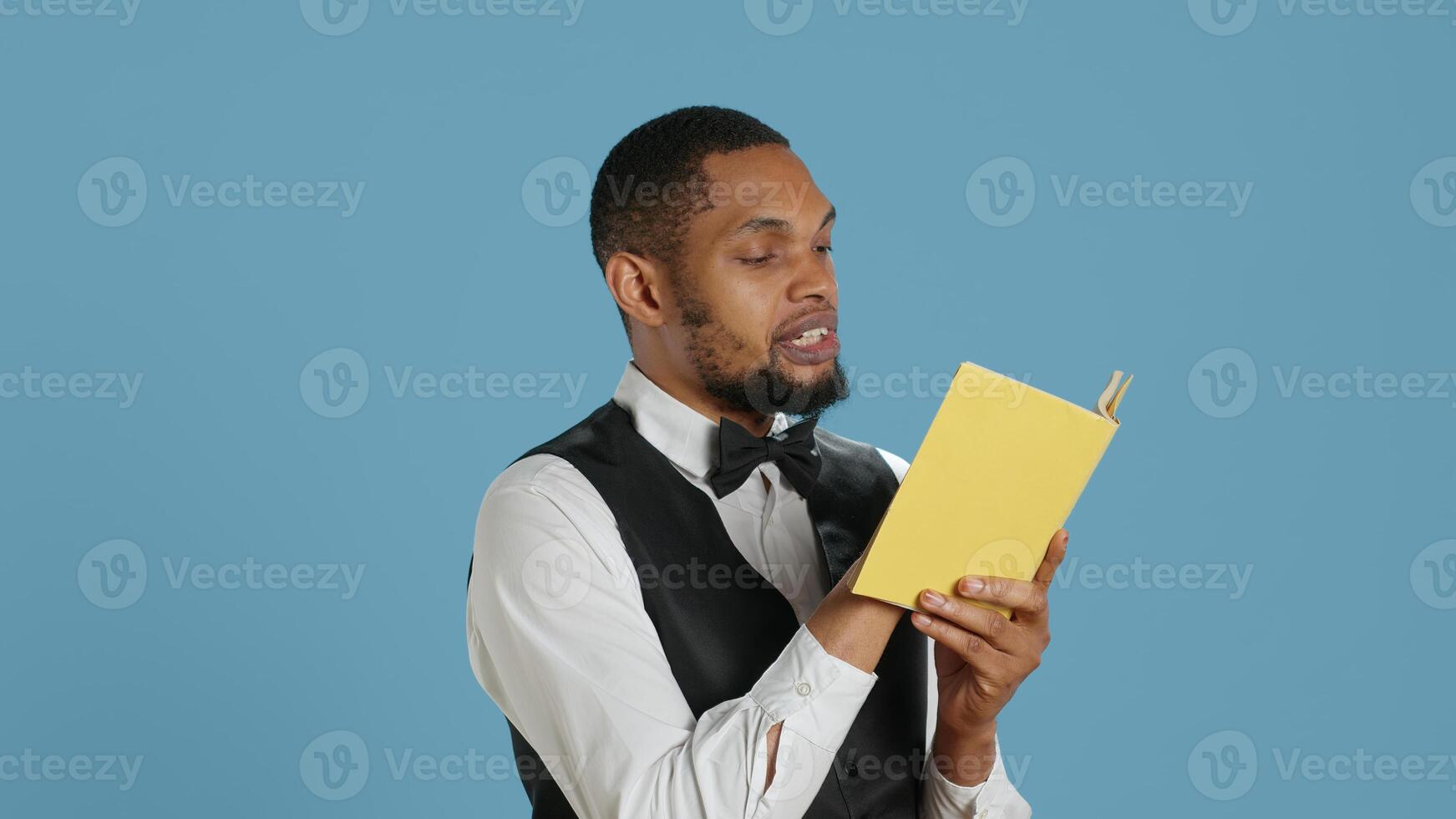 Hotel concierge in tuxedo reading a novel story book in studio, enjoying literature and lecture hobby. Hospitality industry staff working in five star lodging resort, bellhop receptionist. Camera A. photo