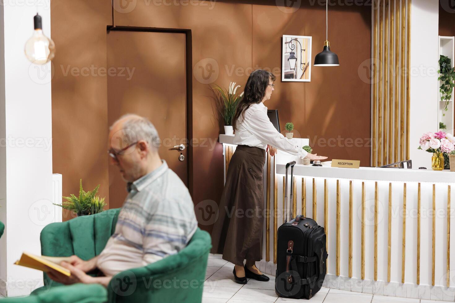 Caucasian elderly woman waiting at the front desk of a contemporary hotel reception requesting lodging. Two senior tourists are waiting in the lounge area which has a couch space for relaxing. photo