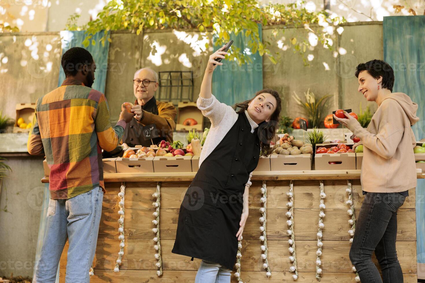 Happy vendor poses for selfie with female customer in front of farmers market stand filled with fresh, locally grown fruits and vegetables. Making use of social media to promote small business market. photo