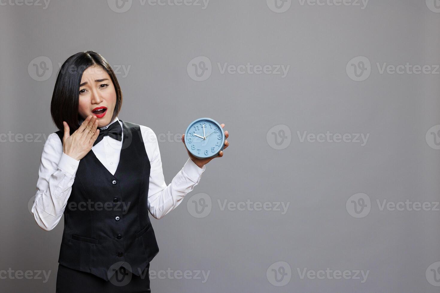 Tired asian woman wearing waitress uniform yawning and covering mouth while holding alarm clock portrait. Sleepy receptionist running late for work, showing time and looking at camera photo
