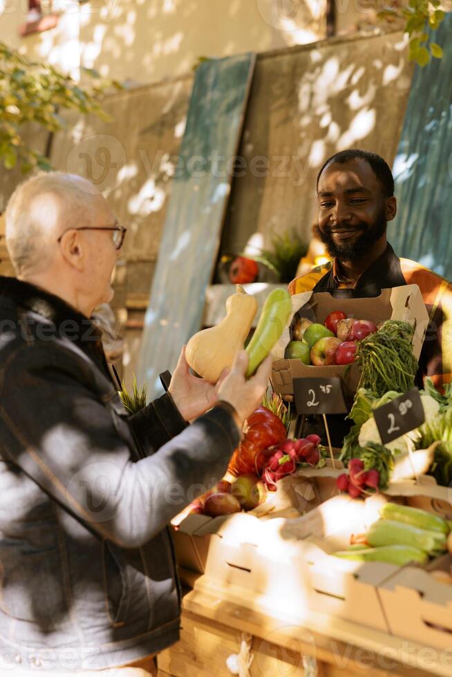 Black male farmer displays organic vegetables from bio garden for elderly customer. Senior client selecting seasonal colorful locally grown fresh produce from greenmarket stand. photo