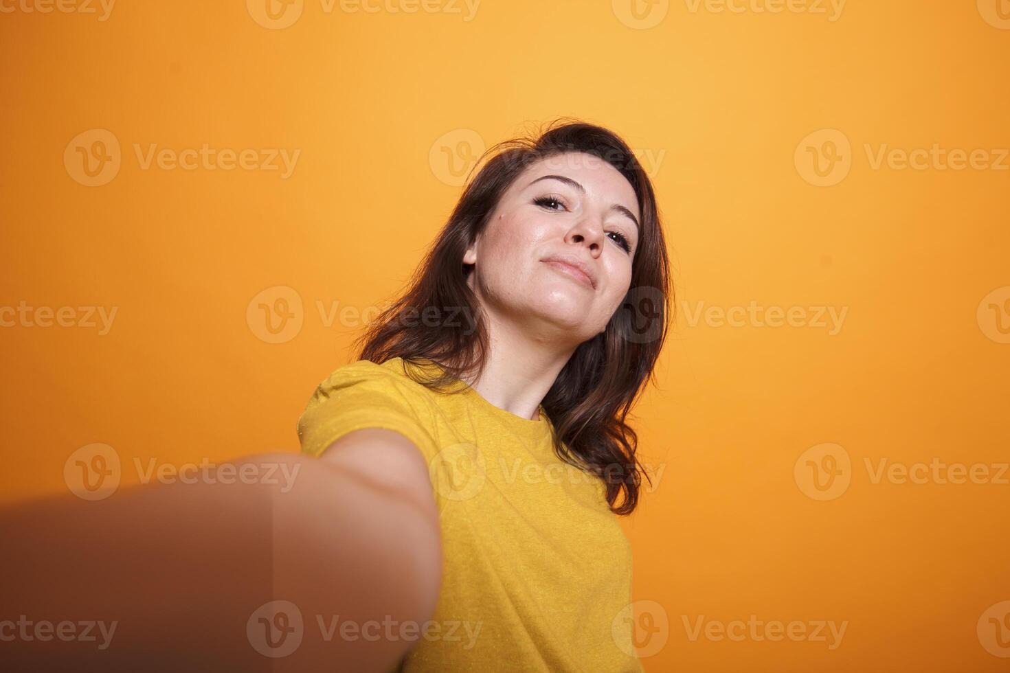 POV of caucasian female individual taking a selfie in studio. Cheerful woman smiling and standing over isolated background, making a personal vlog. Casual person taking picture with smartphone. photo