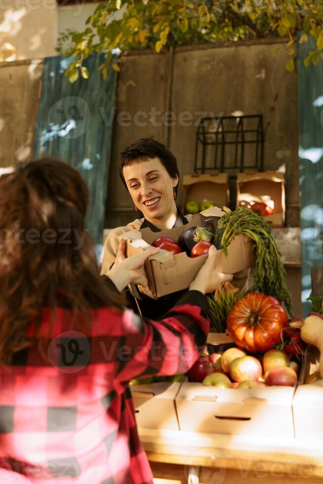 Cheerful, smiling farmer with a box serving customer at farmers market while offering nutritious organic produce. Woman shopping for fresh natural fruits and vegetables at nearby greenmarket. photo