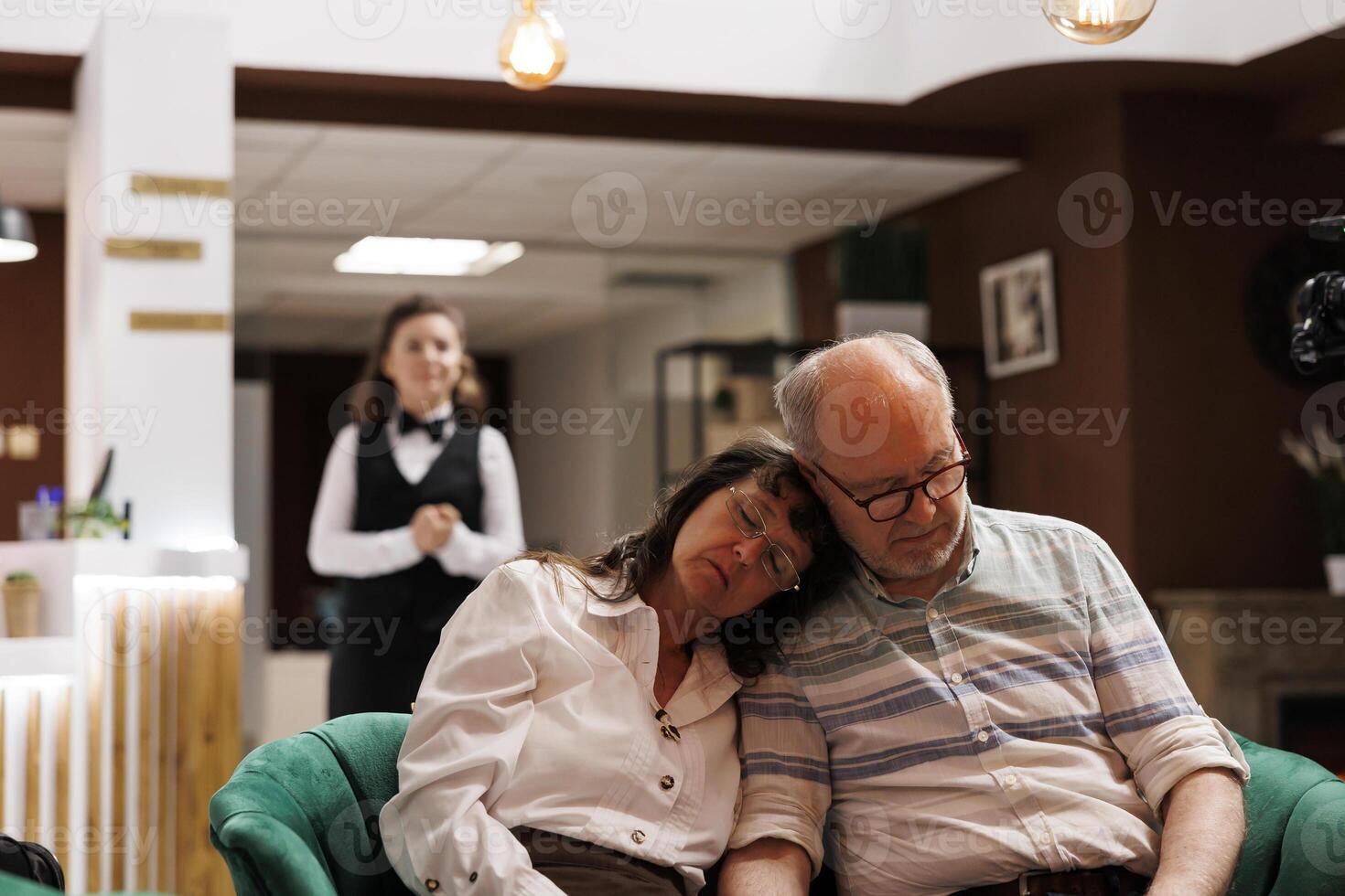 Comfortable hotel lobby with caucasian elderly couple relaxing on comfy couch. Photo showing exhausted retired old man and woman sleeping on sofa in modern lounge area, waiting for check-in.