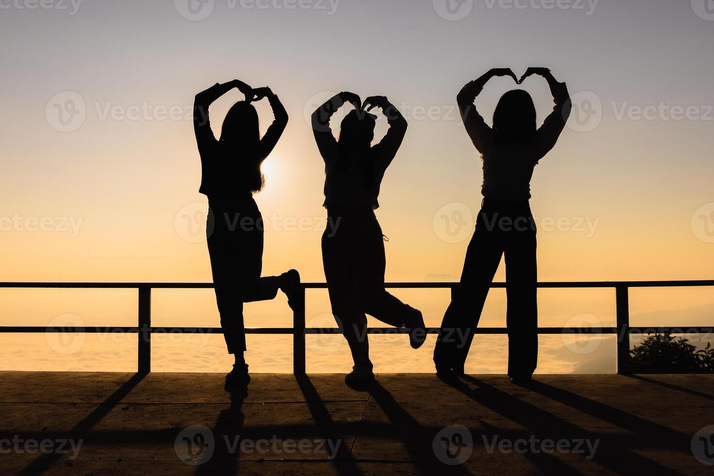 Silhouette of a group of three female tourists raising their hands to form a heart on a mountaintop at sunset. photo