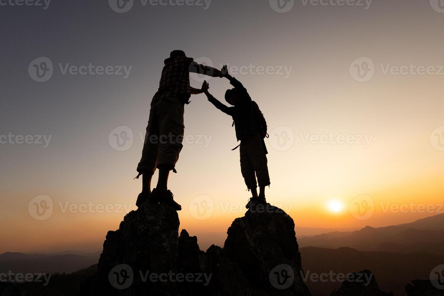 Silhouette of two climbers holding hands on the mountaintop represents success and unity, teamwork, photo