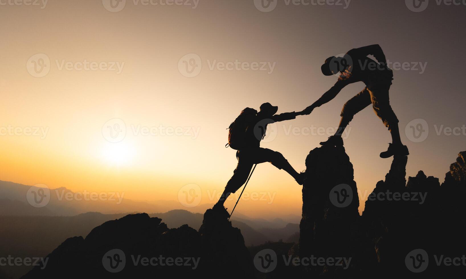 Silhouette of two mountaineers offer helping hand on a rock ridge at sunrise above a valley. Teamwork, unity in work. photo