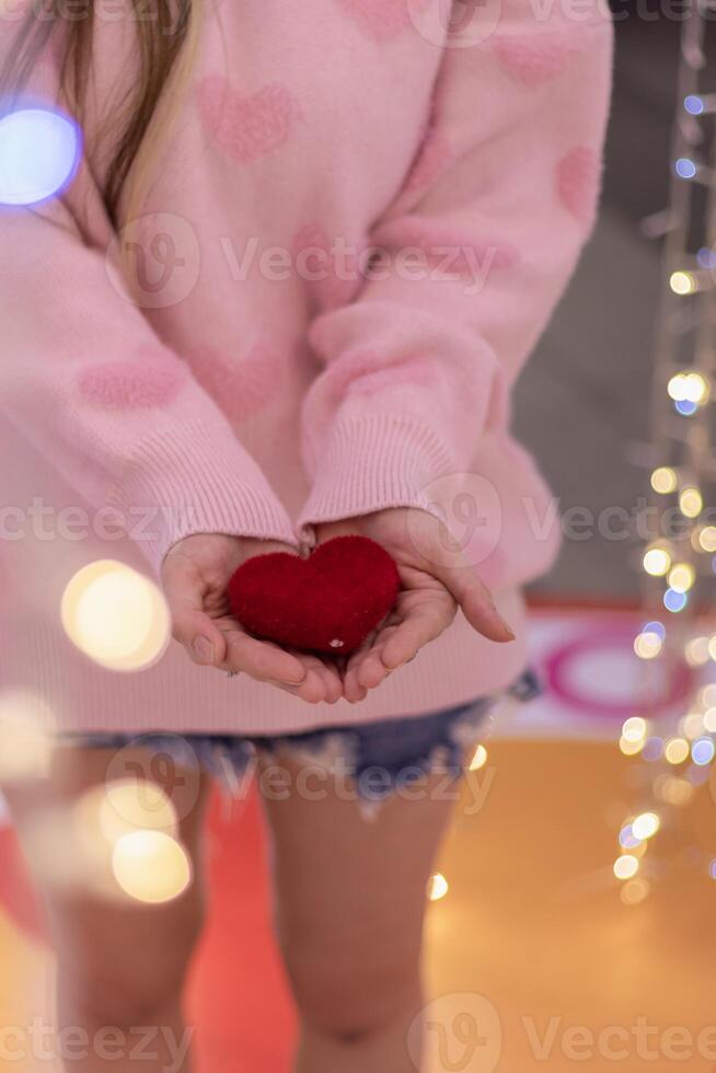 A girl wearing a pink sweater and holding a red heart in her hand on a blurred background of beautiful fairy lights. A woman shows a heart symbol in her hand to tell her boyfriend how she feels. photo