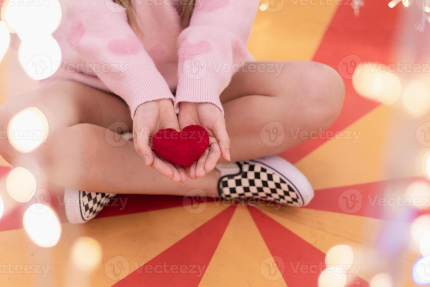 A girl wearing a pink sweater and holding a red heart in her hand on a blurred background of beautiful fairy lights. A woman shows a heart symbol in her hand to tell her boyfriend how she feels. photo