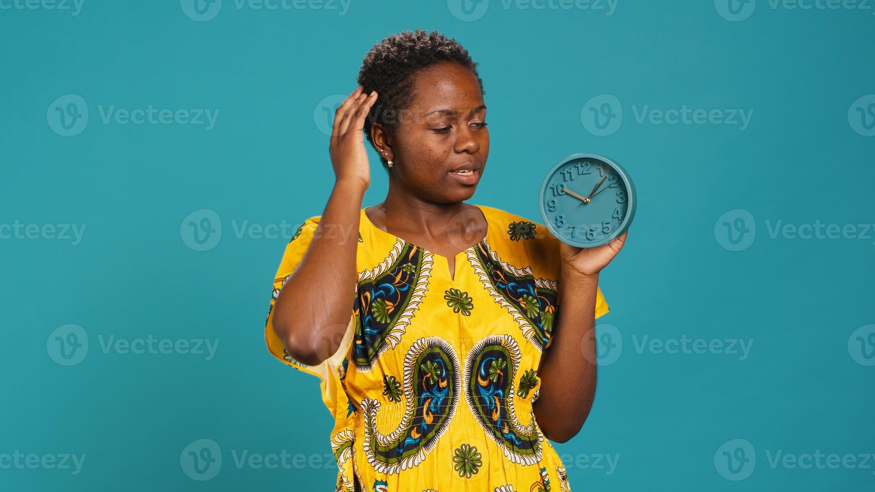 Young woman checking the time on a clock and running late, trying to hurry and stop the delay against blue background. Busy person missing the alarm, having problems with punctuality. Camera A. photo