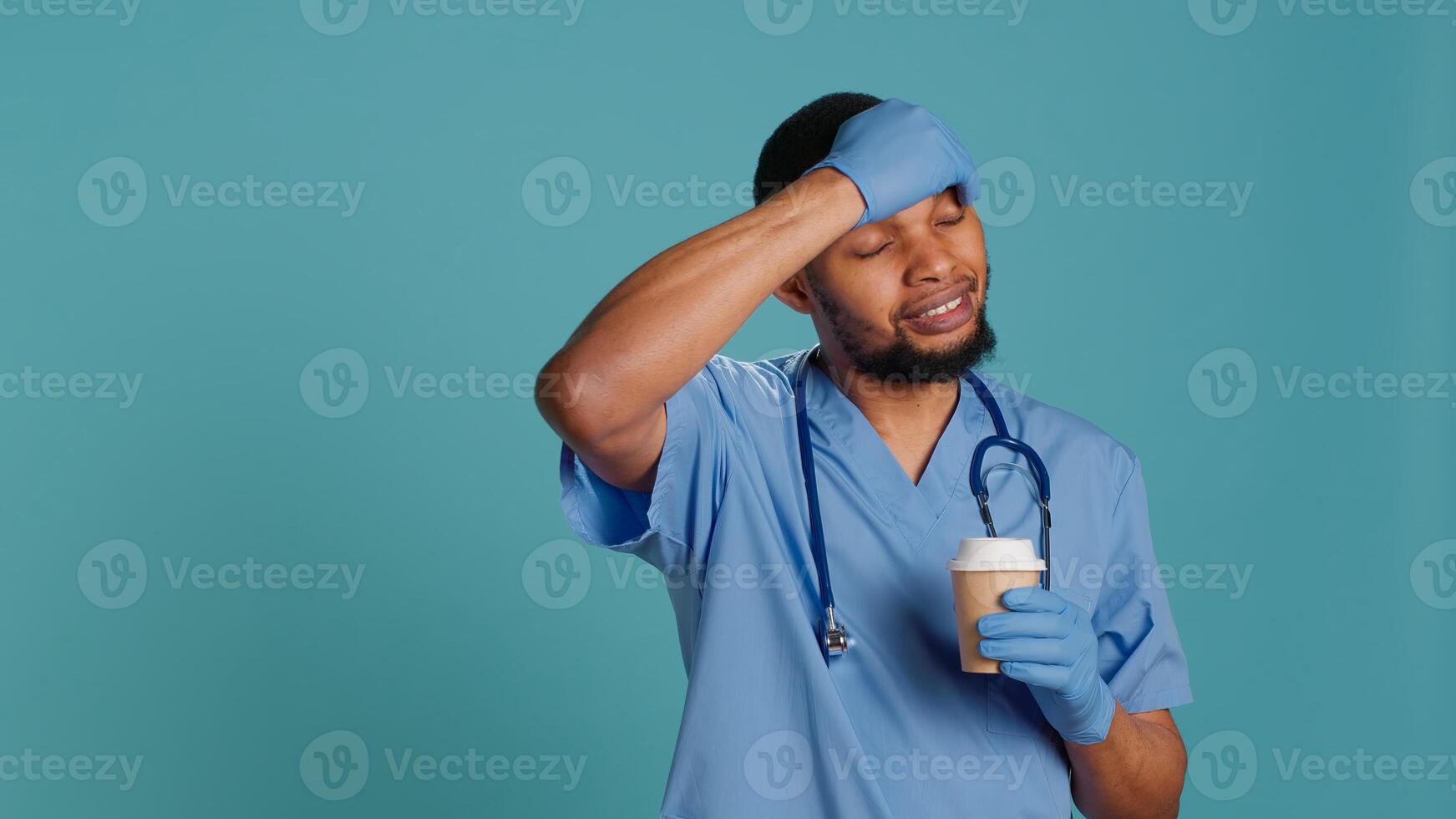 Tired healthcare specialist gaining energy with cup of coffee while at work, enjoying aroma. Fatigued medical practitioner drinking hot beverage, isolated over blue studio background, camera A photo