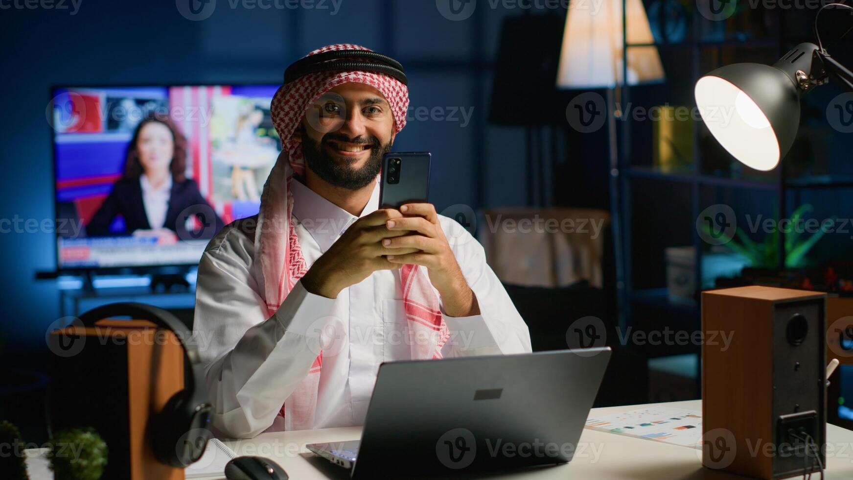 Portrait of smiling Arab freelancer enjoying leisure at home, typing on his smartphone, surfing the net. Happy middle eastern man in traditional attire chatting with friends using mobile phone photo