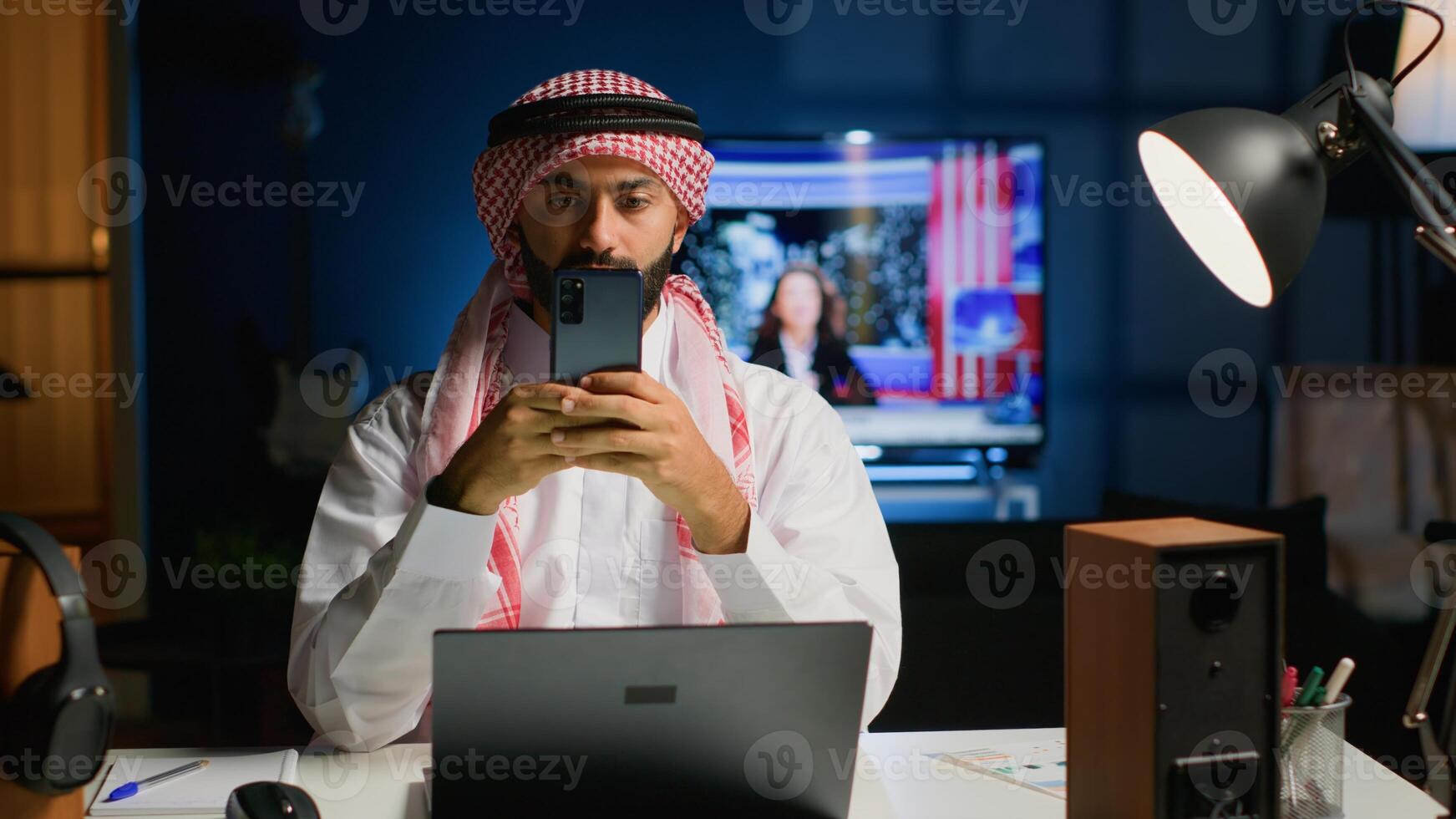 Portrait of smiling Arab freelancer at home, typing on his smartphone, surfing the net. Happy middle eastern man in traditional attire texting his friends using mobile phone photo