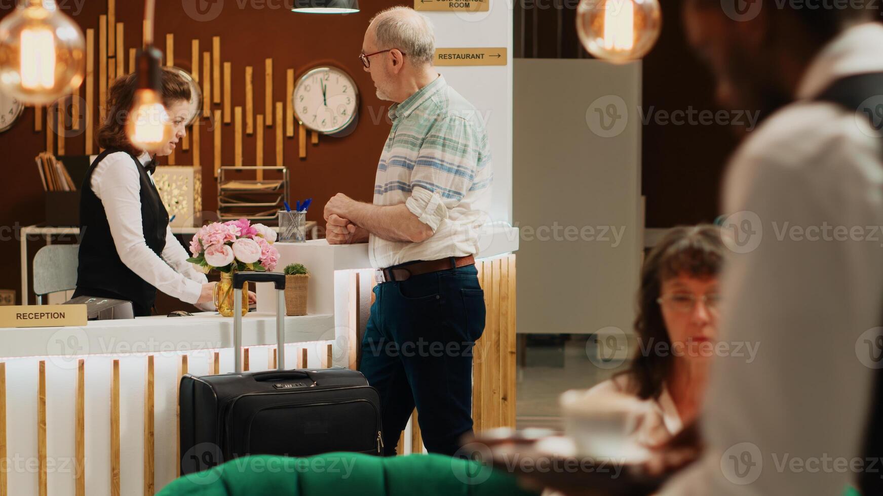 Caucasian man arriving at luxury hotel lobby, goes for check in registration at front desk. Senior traveller heading to front desk with luggage for room booking procedure, retirement age. photo