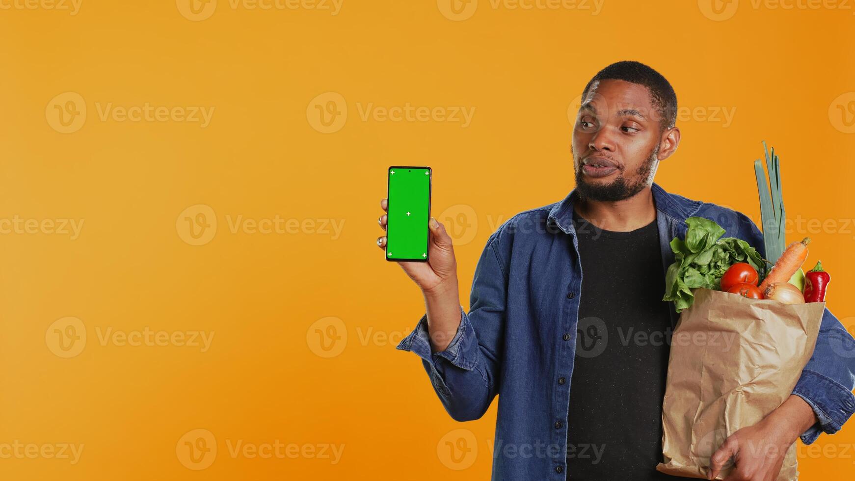 Young guy with a bag of fresh groceries showing greenscreen on his smartphone, presenting an isolated display on phone. Person advertising locally grown produce, zero waste. Camera B. photo