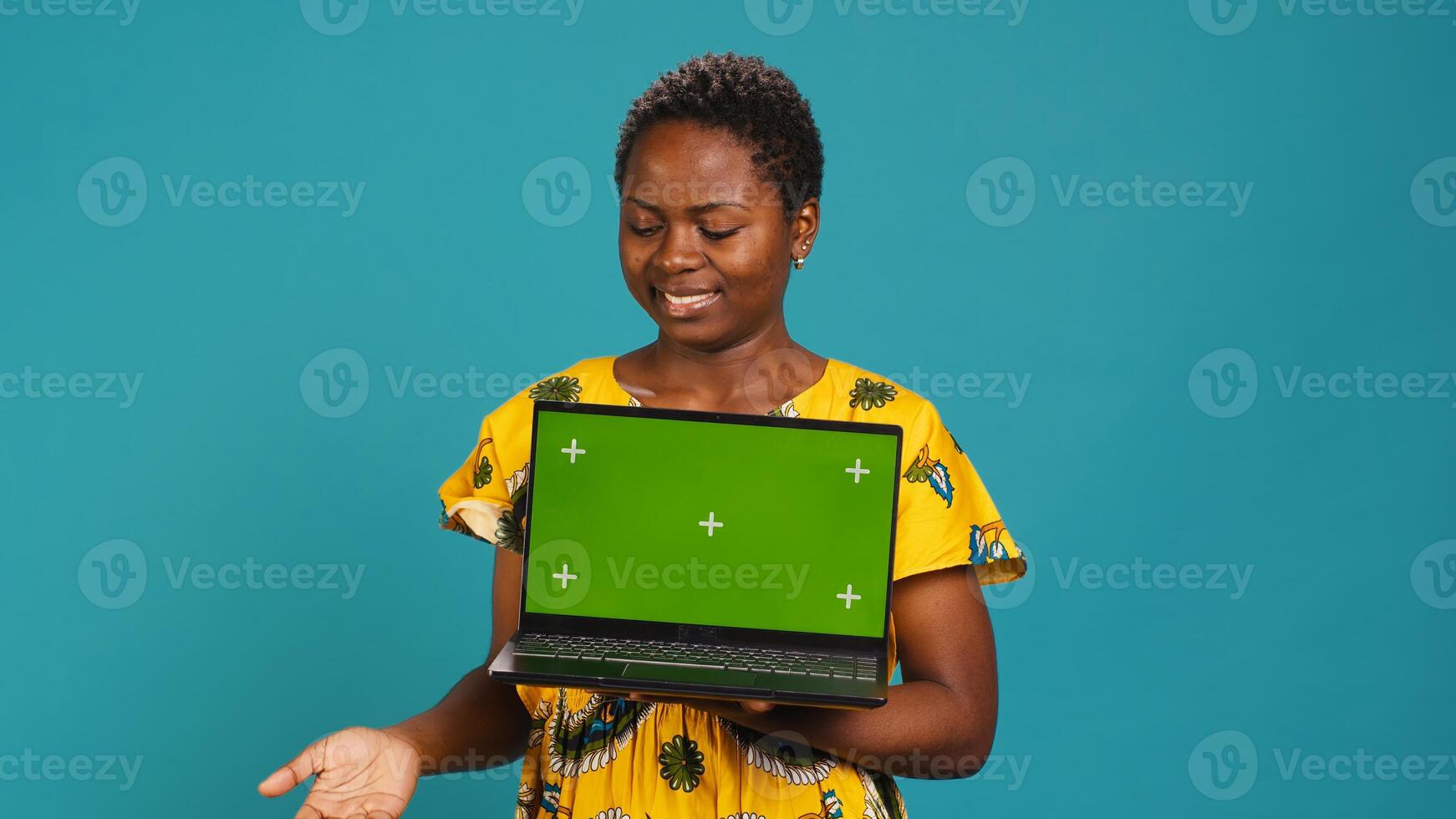 Confident smiling woman presenting a chroma key display on laptop, standing against blue background. Young adult holding a wireless pc and showing a green screen mockup. Camera A. photo