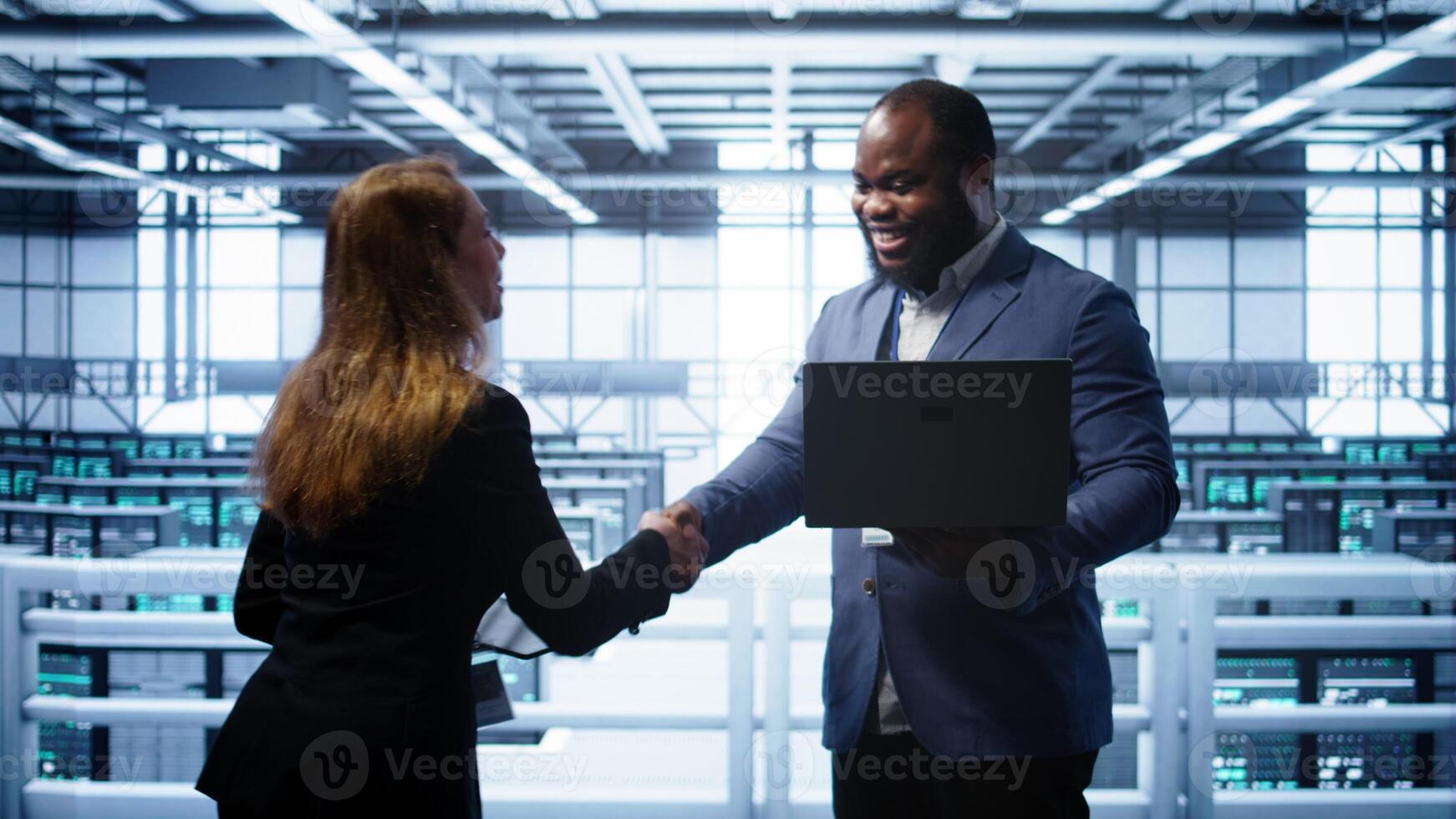 African american admin greeting inspector arriving in data center to supervise operations. IT expert in server hub workspace doing hand shake with supervisor monitoring performance, camera B photo