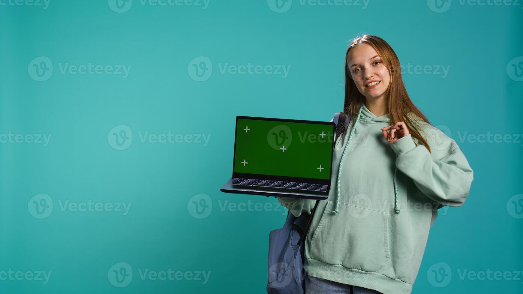 Happy girl presenting laptop with green screen display, isolated over studio backdrop. Radiant teenager creating promoting advertisement with mockup notebook device, camera B photo