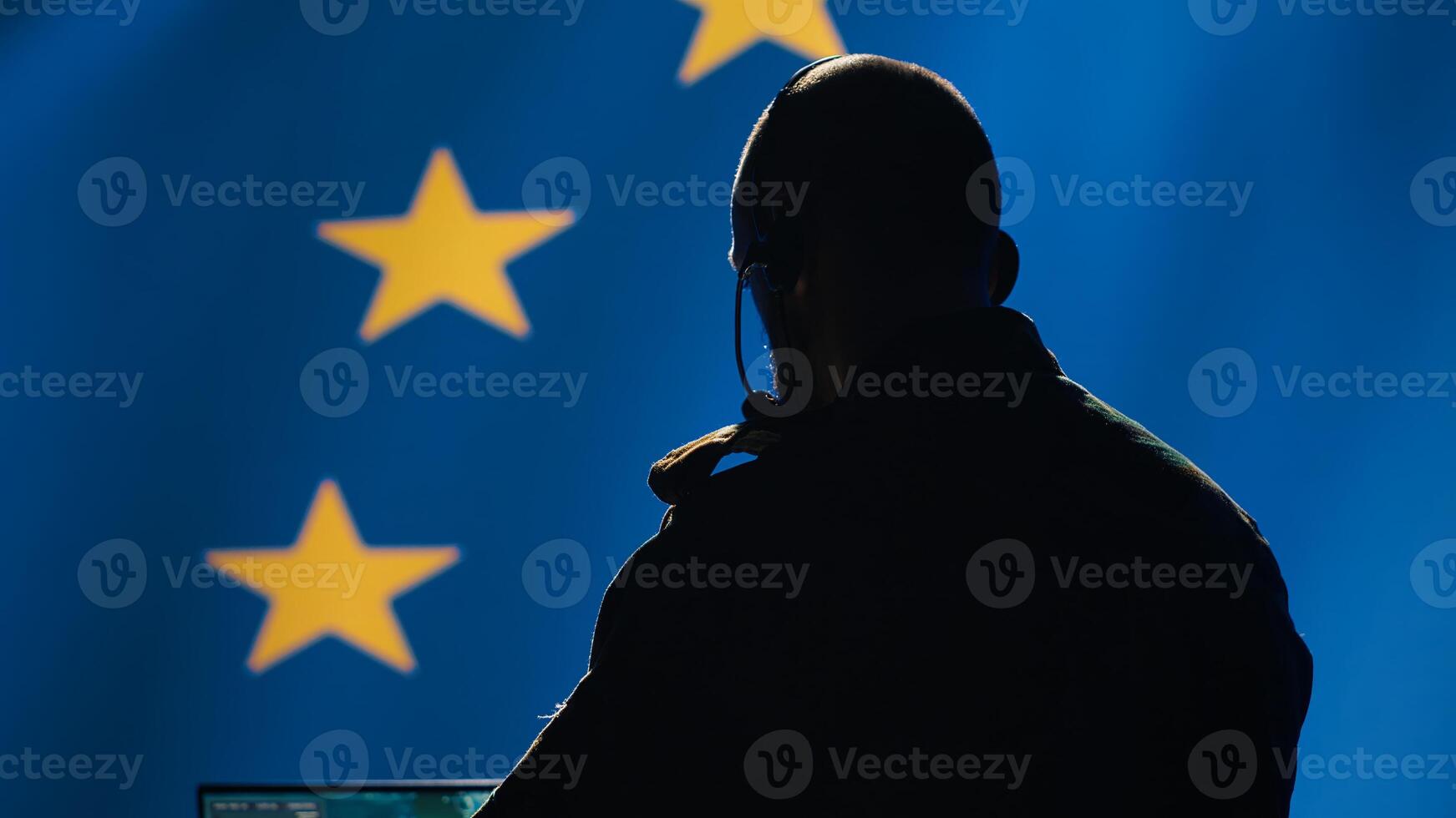European Union military dispatcher looking at surveillance broadcast, relaying informations for troops using headset. Telecommunications operator over EU flag background, coordinating troops, camera A photo