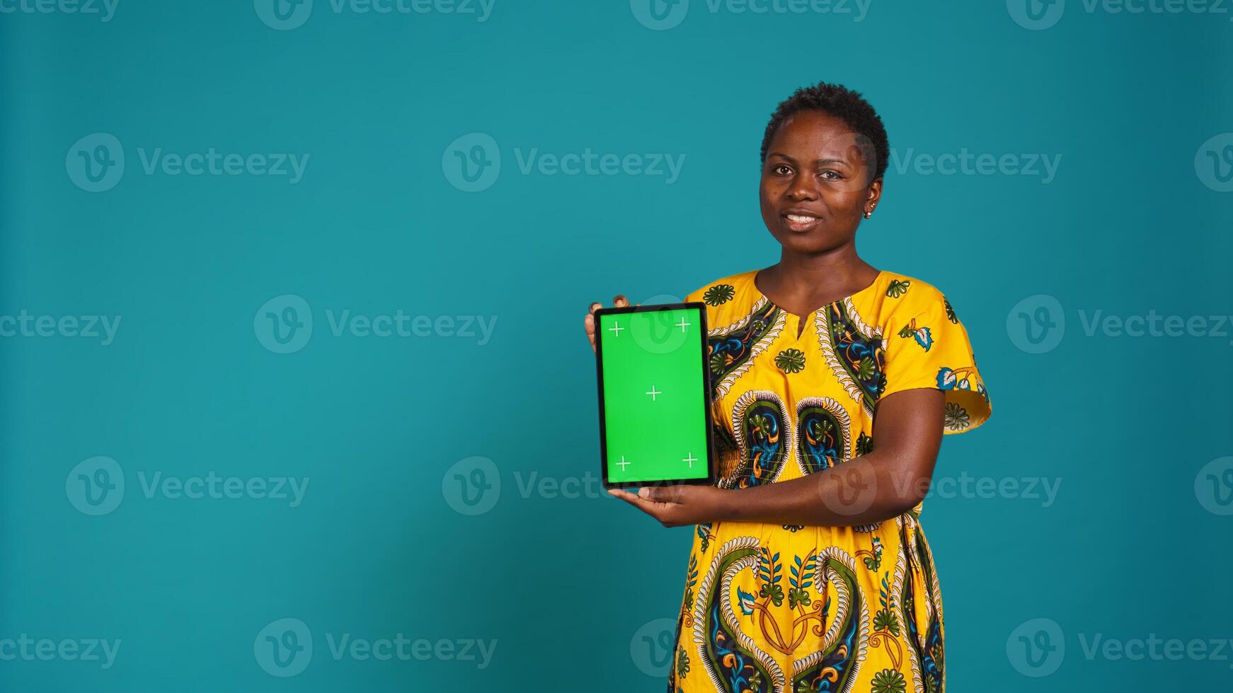 Feminine person in native clothing holds a tablet with mockup screen, creating an advertisement against blue background in studio. Young woman smiling and presenting copy space. Camera B. photo