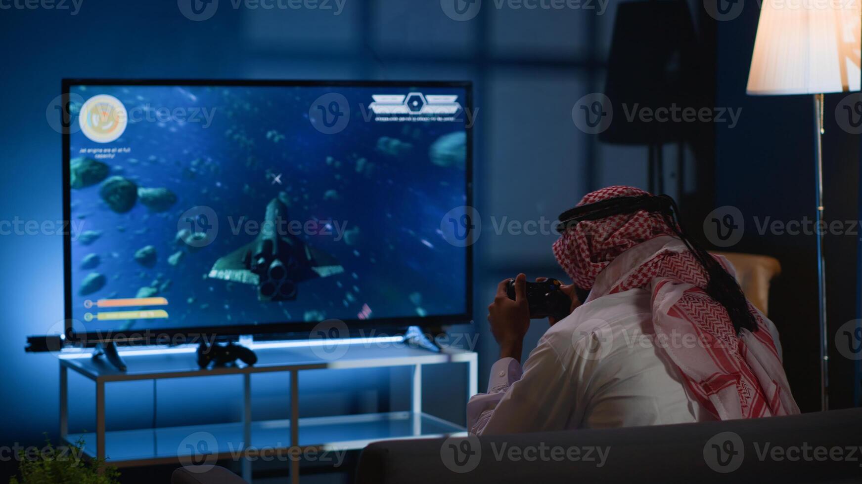 Arabic gamer playing classic arcade action videogame, flying through space debris. Muslim man enjoying leisure time at home using high tech gaming console to solve missions in singleplayer game photo