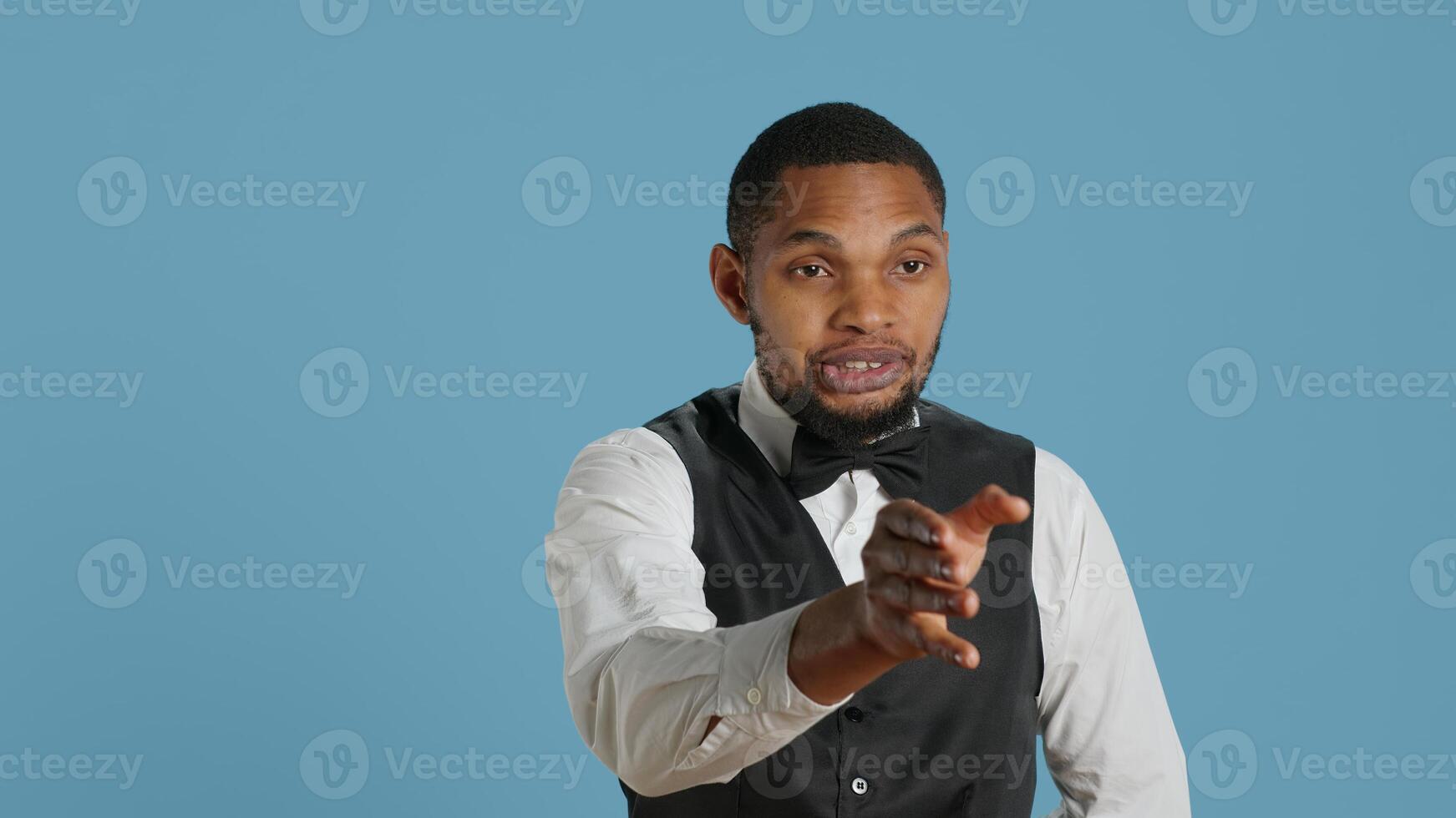 Bellboy hotel porter welcoming guests at the five star lodging resort, smiling and greeting people against blue background. Employee wearing a tuxedo and tie, tourism industry. Camera A. photo