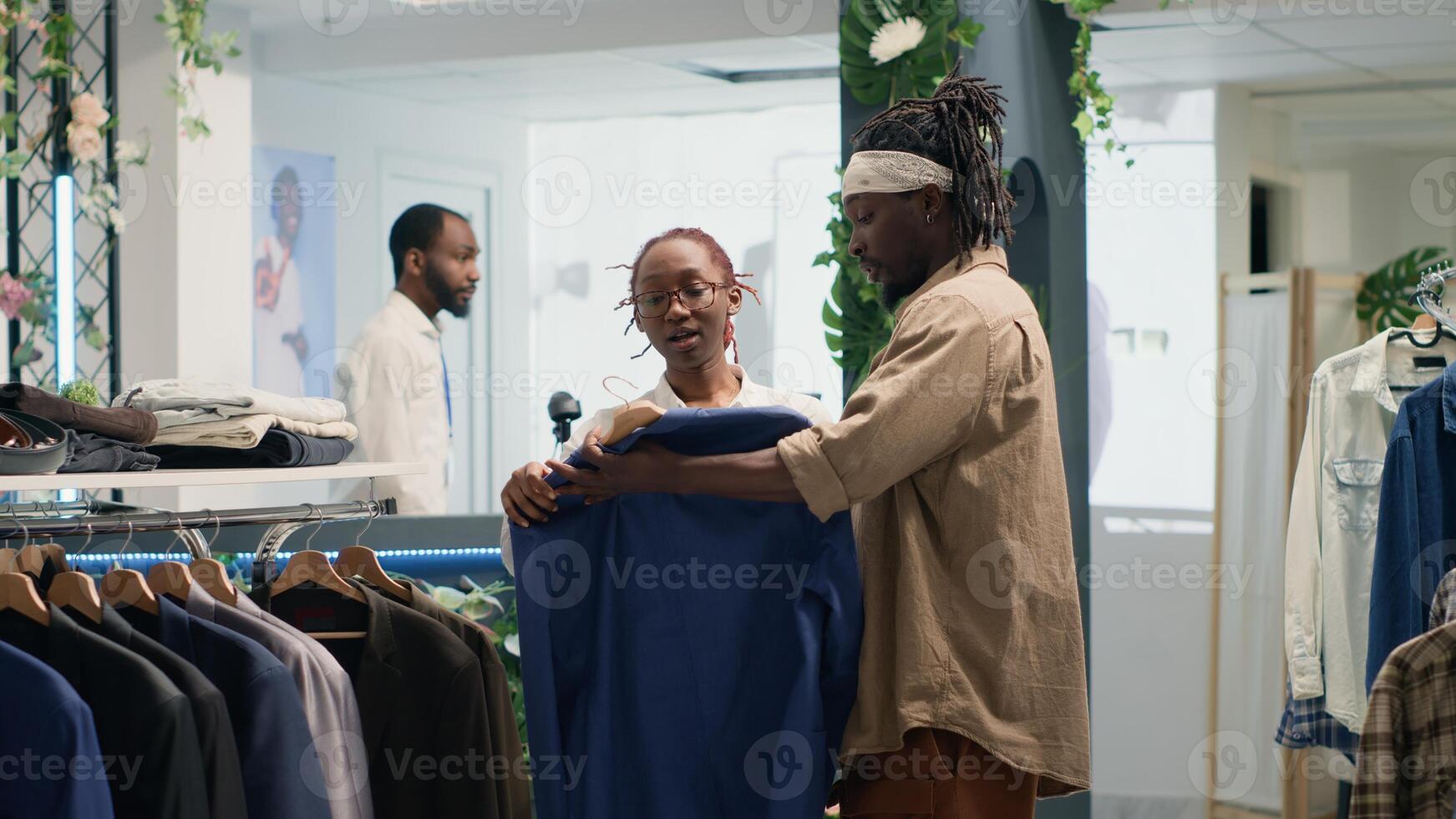 Man doing shopping in elegant fashion boutique, assisted by friendly shopkeeper with clothes. Customer receiving help from woman while trying on blazer from new collection stock photo